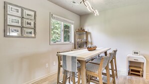 Un comedor con mesa de madera, sillas, un tapete, una ventana con persianas, una repisa con vasos y un cuadro en la pared.