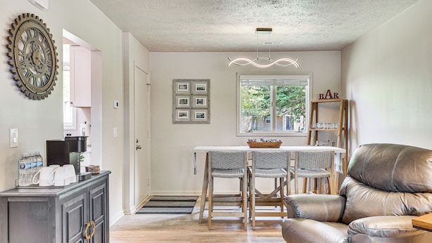 Un comedor con una mesa y sillas, un sofá de cuero, un reloj decorativo en la pared y una ventana con vistas a árboles.