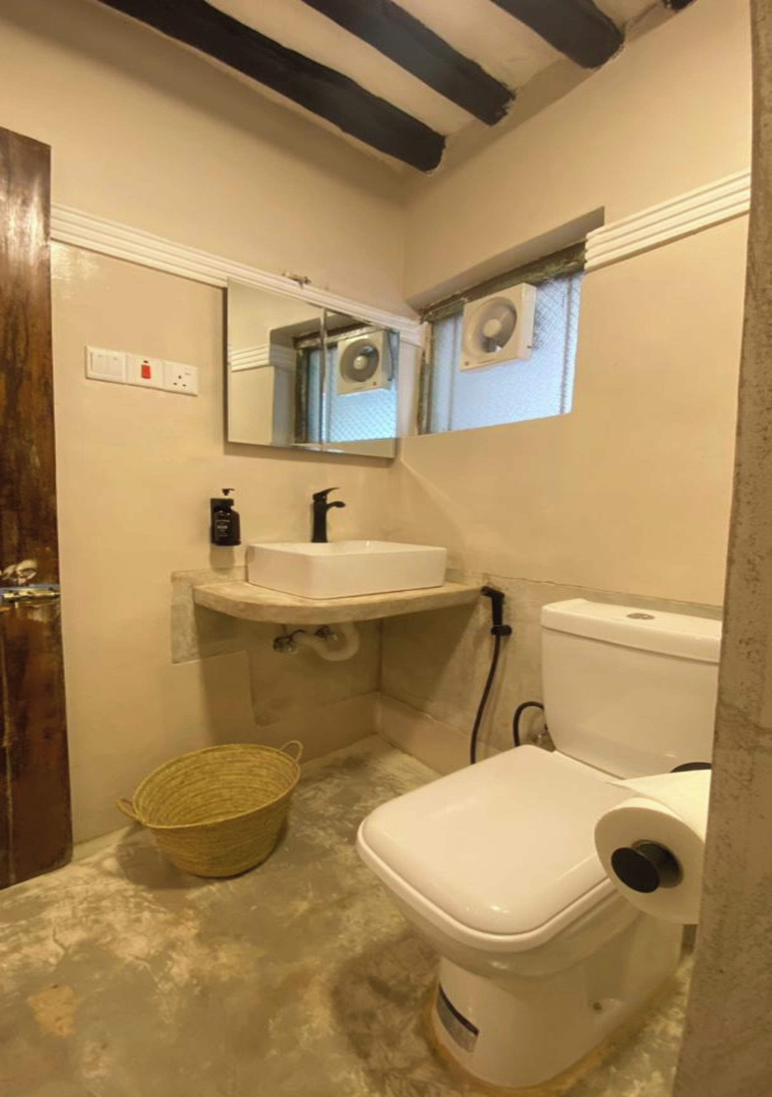 A small bathroom with a white sink, a toilet, and a mirror. There is a basket on the floor and a window above the toilet.