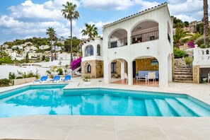 Área ao lado da piscina, com piscina azul cristalina, edifício branco com arcos e vista para o morro.