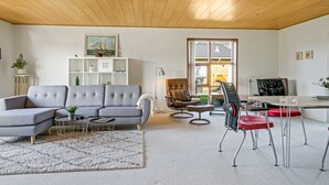 Uma sala de estar moderna com um sofá seccional cinza, uma mesa de centro redonda, uma mesa de jantar com cadeiras vermelhas e uma grande janela com vista para o exterior.