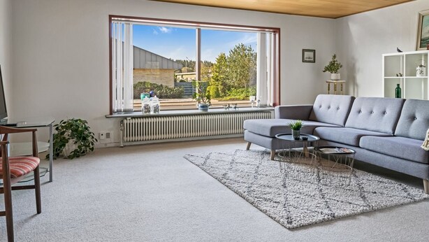 Uma sala de estar moderna com um sofá cinzento, uma mesa de centro redonda e uma janela ampla com vista para um edifício e árvores.
