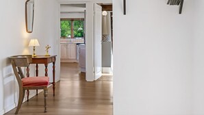 Um corredor com uma cadeira de madeira, uma mesa com abajur e um espelho. É possível ver uma cozinha ao fundo, com uma janela que mostra áreas verdes lá fora.