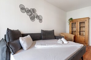 Ein ordentlich bezogenes Bett mit Kissen, ein graues Sofa, ein Holzschrank und ein Wandbild.