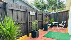 Un patio trasero con cerca de madera, plantas en macetas, un pequeño área de patio con mesa y sillas, y un tapete verde.