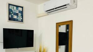 Una habitación con televisión de pantalla plana, un escritorio blanco, un florero azul turquesa con plantas secas, un espejo con marco de madera y un equipo de aire acondicionado.