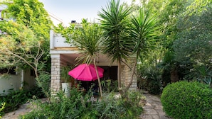 Een stenen pad dat leidt naar een gebouw met een roze parasol, omgeven door weelderige begroeiing en verschillende planten.