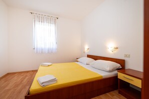A single-bed room with a wooden headboard, a bedside table with a drawer, a window with sheer curtains, and wall-mounted lights.