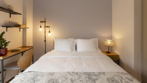 Habitación de hotel ordenada con cama, mesita de noche, lámpara y escritorio con silla y planta en maceta.