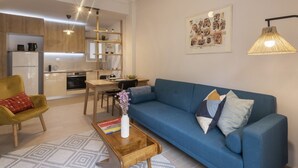 Un salon moderne avec un canapé bleu, une table basse en bois et un fauteuil jaune. On aperçoit une cuisine à l’arrière-plan, avec des meubles de rangement en bois et un coin repas.