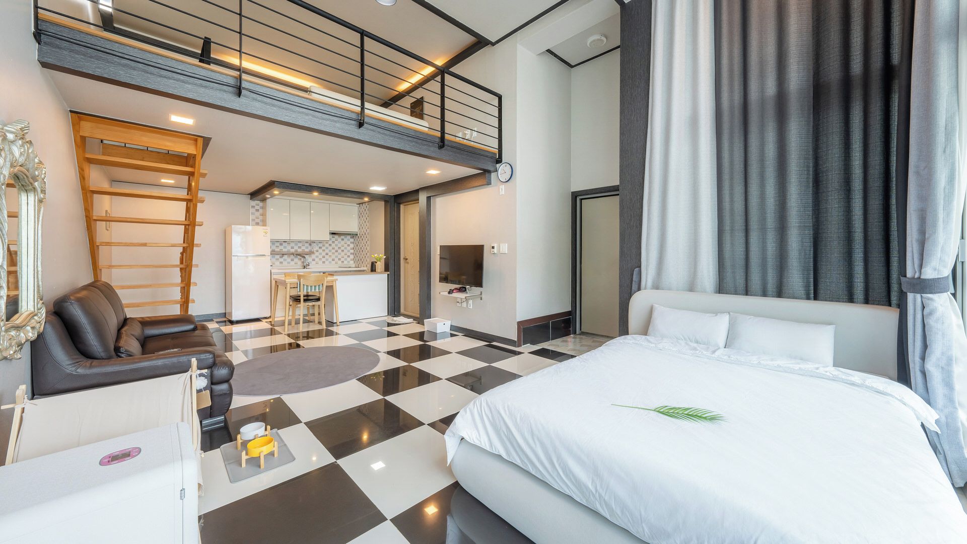 Habitación de hotel con piso a cuadros, una cama con ropa de cama blanca, un escritorio con una silla, una kitchenette y un balcón con barandilla.