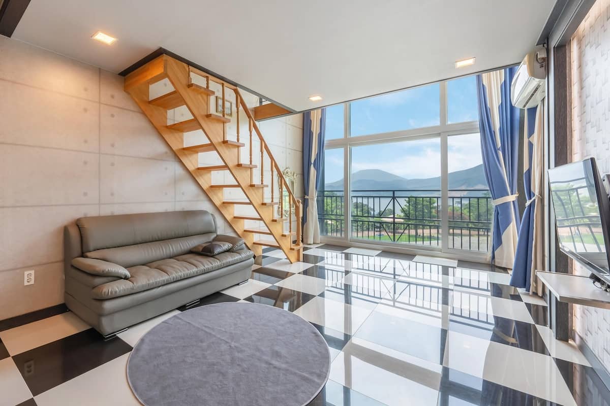 Ein modernes Wohnzimmer mit grauem Sofa, einer Holztreppe und großen Fenstern mit Blick ins Freie.