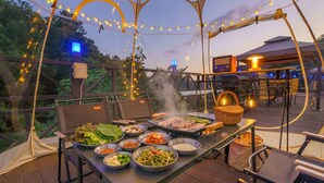 Un área de comedor al aire libre con una parrilla, diversos platos y luces colgantes.
