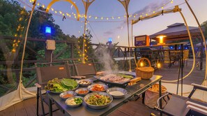 Un área de comedor al aire libre con una parrilla, diversos platos y luces colgantes.