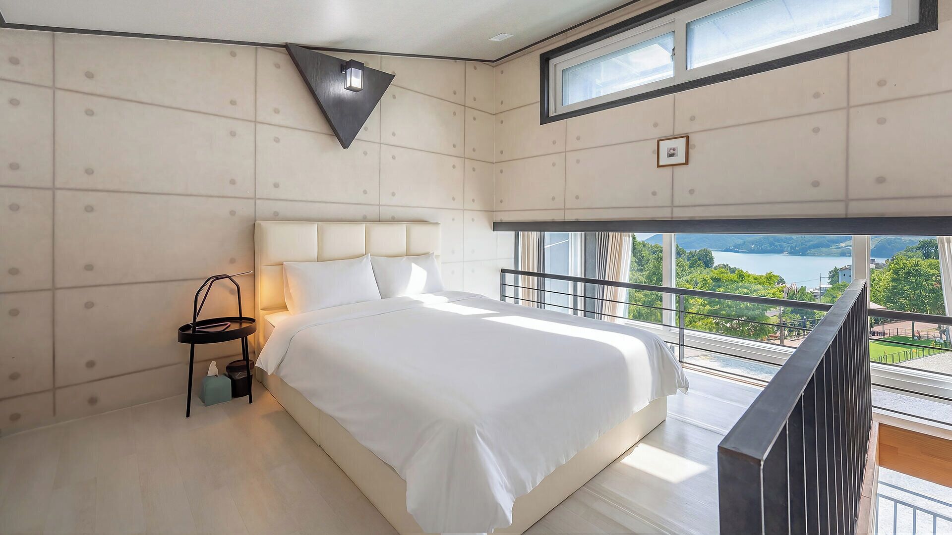 A modern bedroom with a large bed, a nightstand, a wall-mounted light, and a window with a view of a lake.