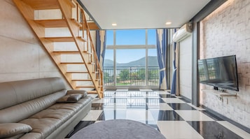 A modern living room with a sofa, a TV, a staircase, and a large window offering a view of the outdoors.