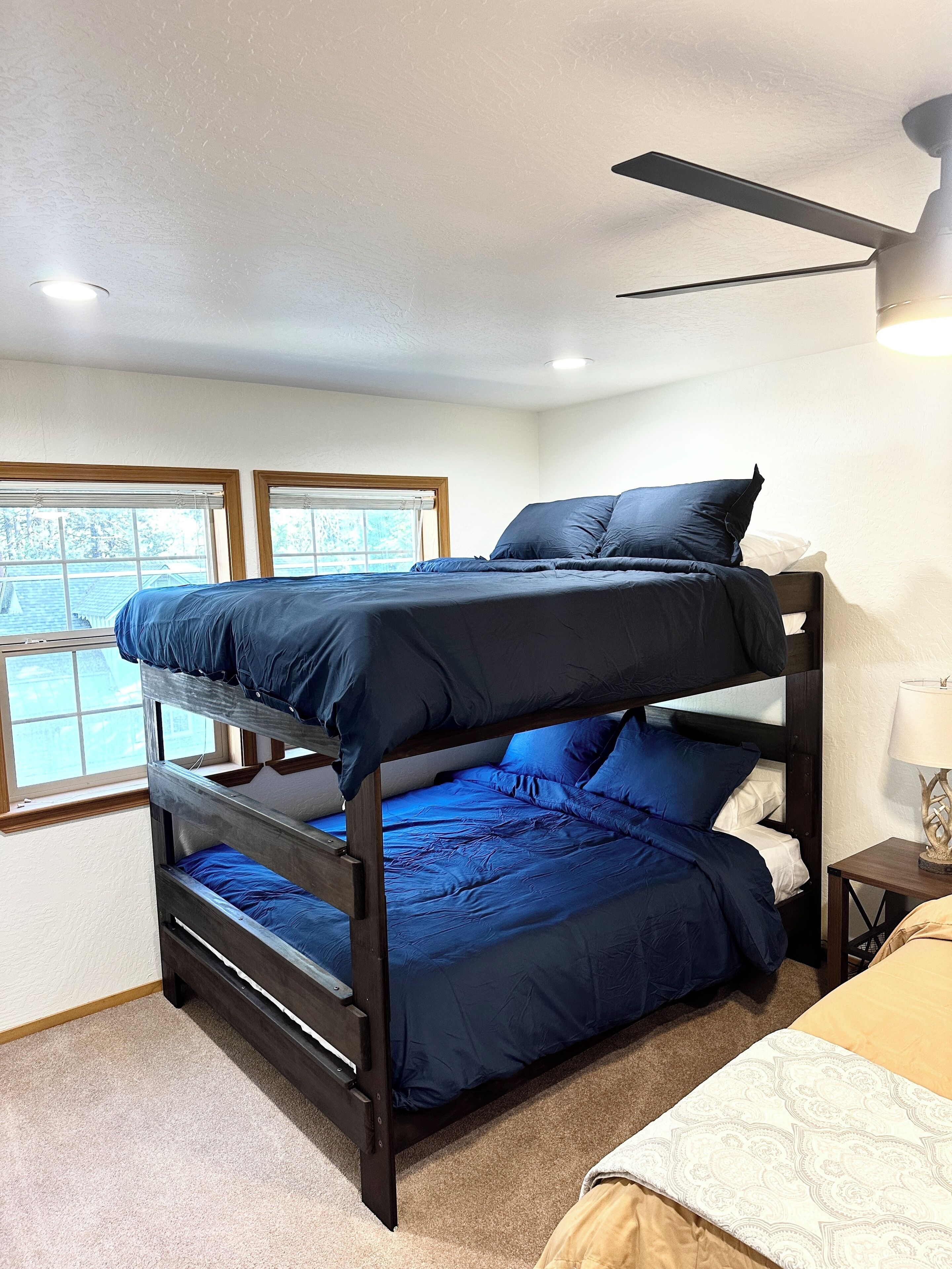 A bunk bed room with a ceiling fan, a window with blinds, and a lamp on a bedside table.