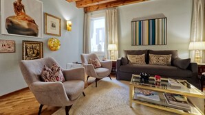 Een woonkamer met houten plafond, een bank, twee fauteuils, een salontafel, en diverse wanddecoraties, waaronder ingelijste foto's en een schilderij.