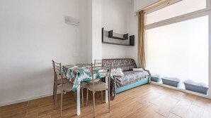 Una habitación con una mesa de comedor, sillas, un sofá, una ventana con cortinas y un estante fijado a la pared.