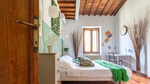 Een slaapkamer met een houten vloer, een bed met een groene deken, een bureau, een stoel en een raam.