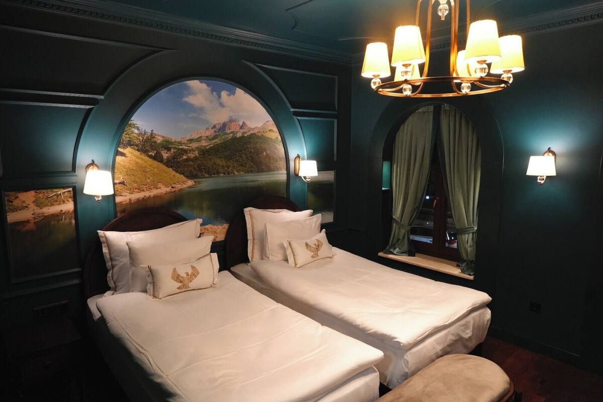 Una habitación de hotel con dos camas, una araña y un mural pintoresco de una montaña y un lago.