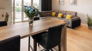 Ein modernes Wohnzimmer mit einem Holzesstisch, dunklen Stühlen, einem grauen Sofa mit gelben Kissen und einem großen Fenster mit Blick auf die Stadt.
