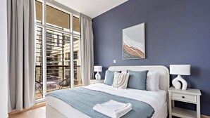 Un dormitorio moderno con una cama grande, dos mesitas de noche, una ventana con cortinas y vistas a un edificio.