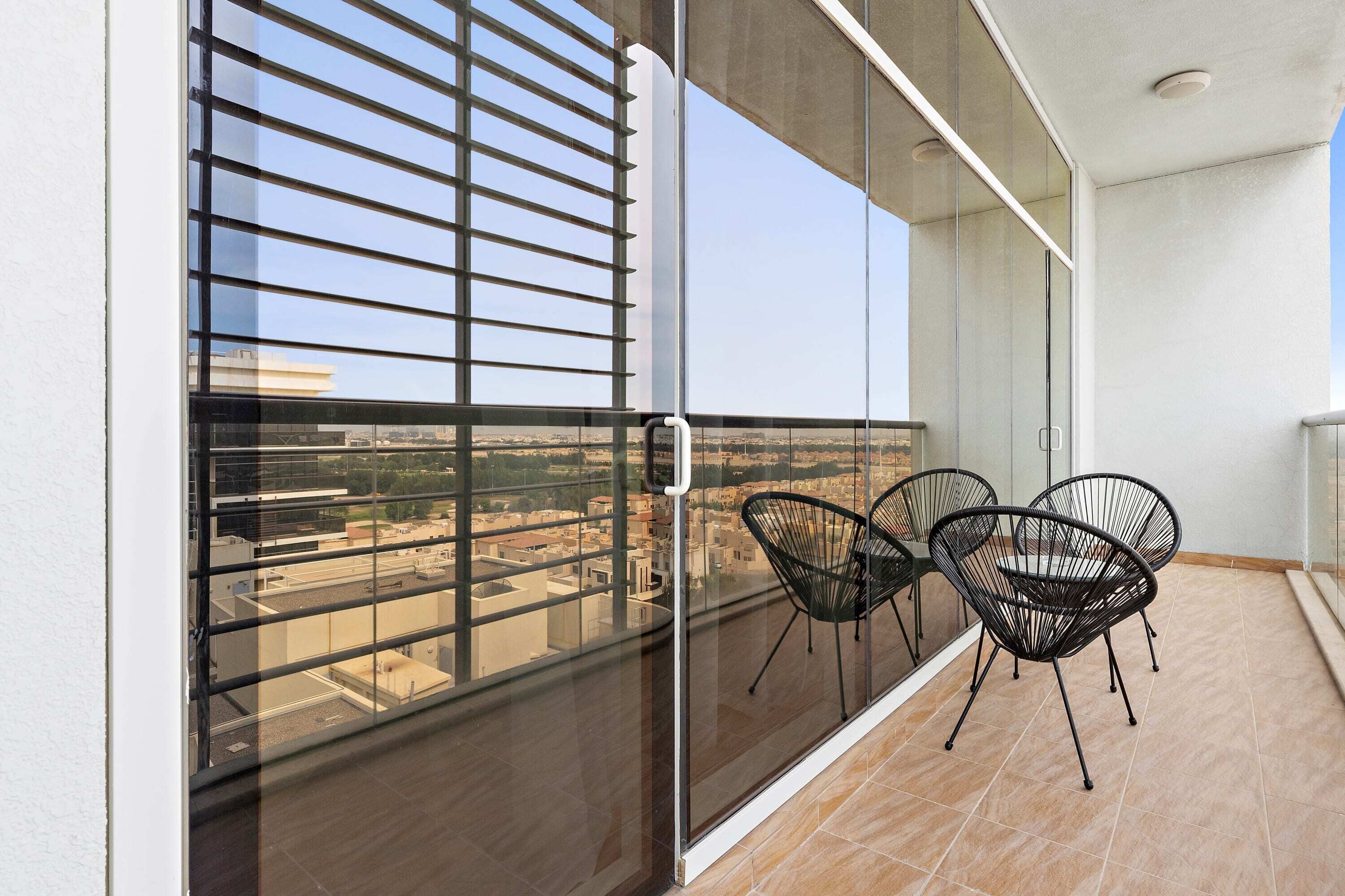 Un balcon avec une porte vitrée, deux chaises noires et une vue sur des immeubles et un ciel dégagé.