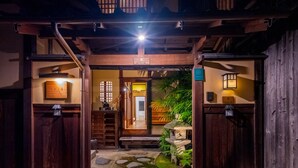 Una entrada japonesa tradicional con puertas de madera, un sendero de piedra y un pequeño jardín con rocas y plantas.