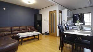 A living room with a brown leather sofa, a dining table with chairs, a small table with a vase, and a kitchen area in the background.