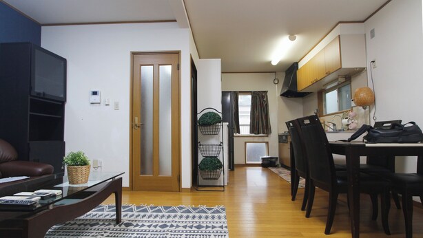 A living room with a sofa, coffee table, and a patterned rug. There is a door leading to a kitchen with a dining table and chairs.