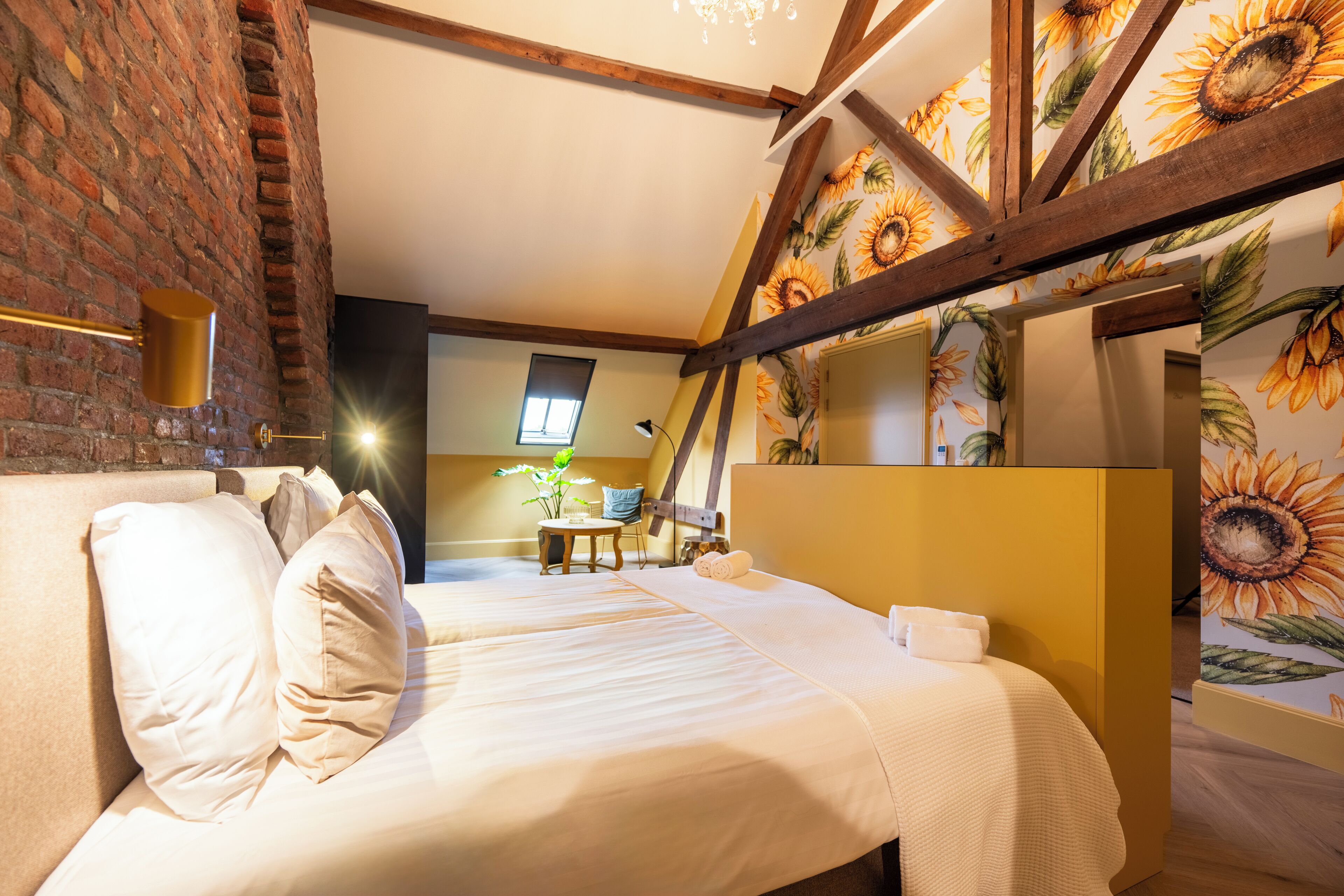 Ein Zimmer mit Backsteinwand, Holzbalken und einem Sonnenblumen-Wandbild. Es gibt ein Bett mit weißer Bettwäsche sowie einen kleinen Tisch mit einem Stuhl.