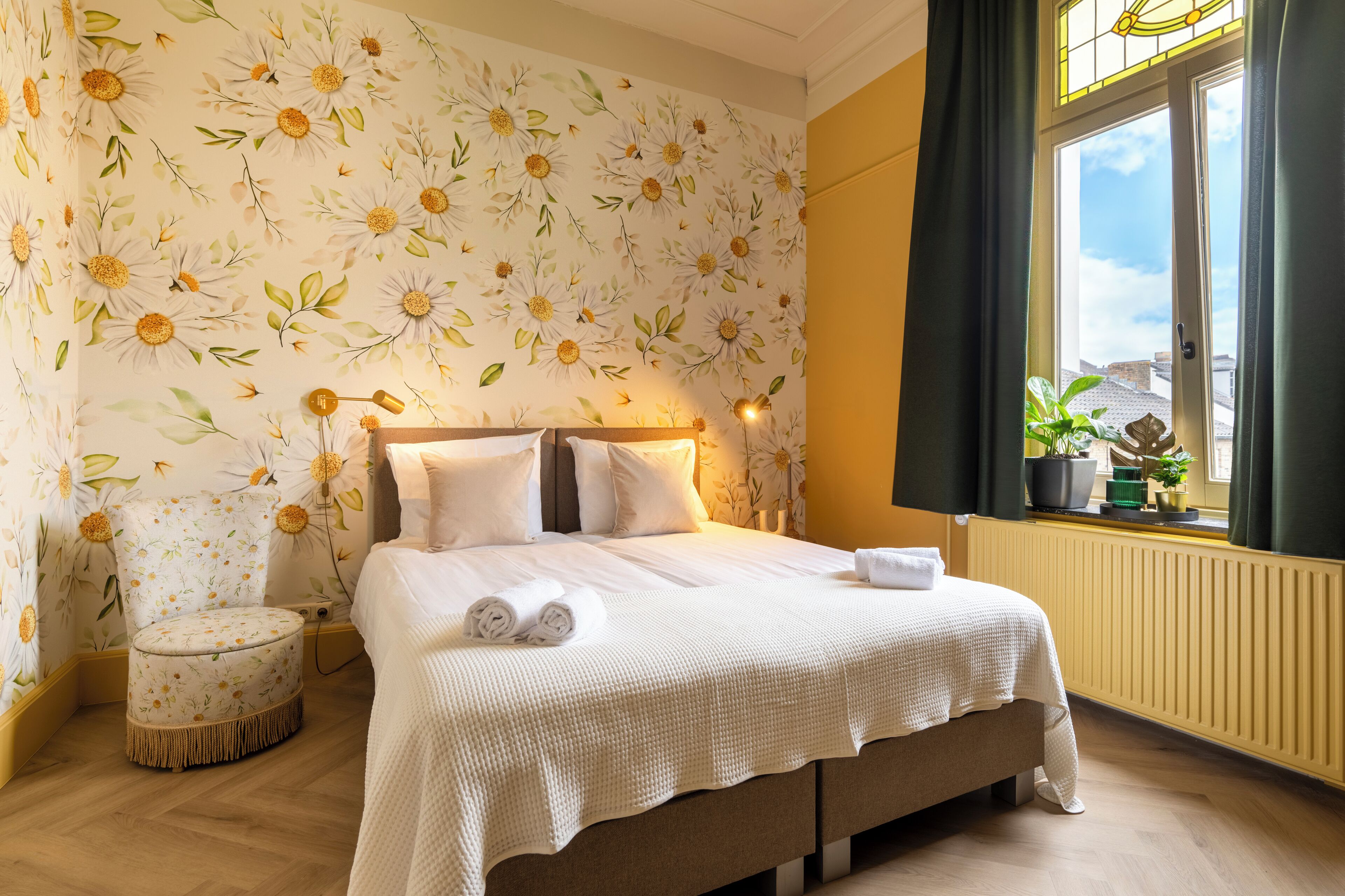 A bedroom with a floral wallpaper, a bed with white linens, a round ottoman, and a window with curtains.