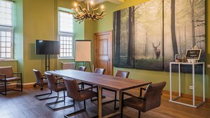 Uma sala de conferência com uma mesa comprida, cadeiras, um lustre, um quadro de uma floresta, uma lousa branca e uma televisão.