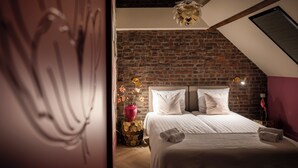 Quarto com parede de tijolos, cama com roupa de cama branca, vaso vermelho com flores e um lustre de teto exclusivo.
