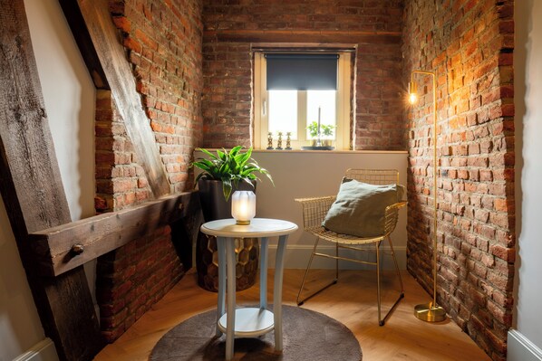 A cozy room with exposed brick walls, a small round table with a lamp, a chair, and a window with plants.