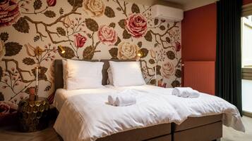 Una cama bien tendida con sábanas blancas y dos almohadas, una pared decorativa con papel tapiz floral, una lámpara de noche y un radiador.