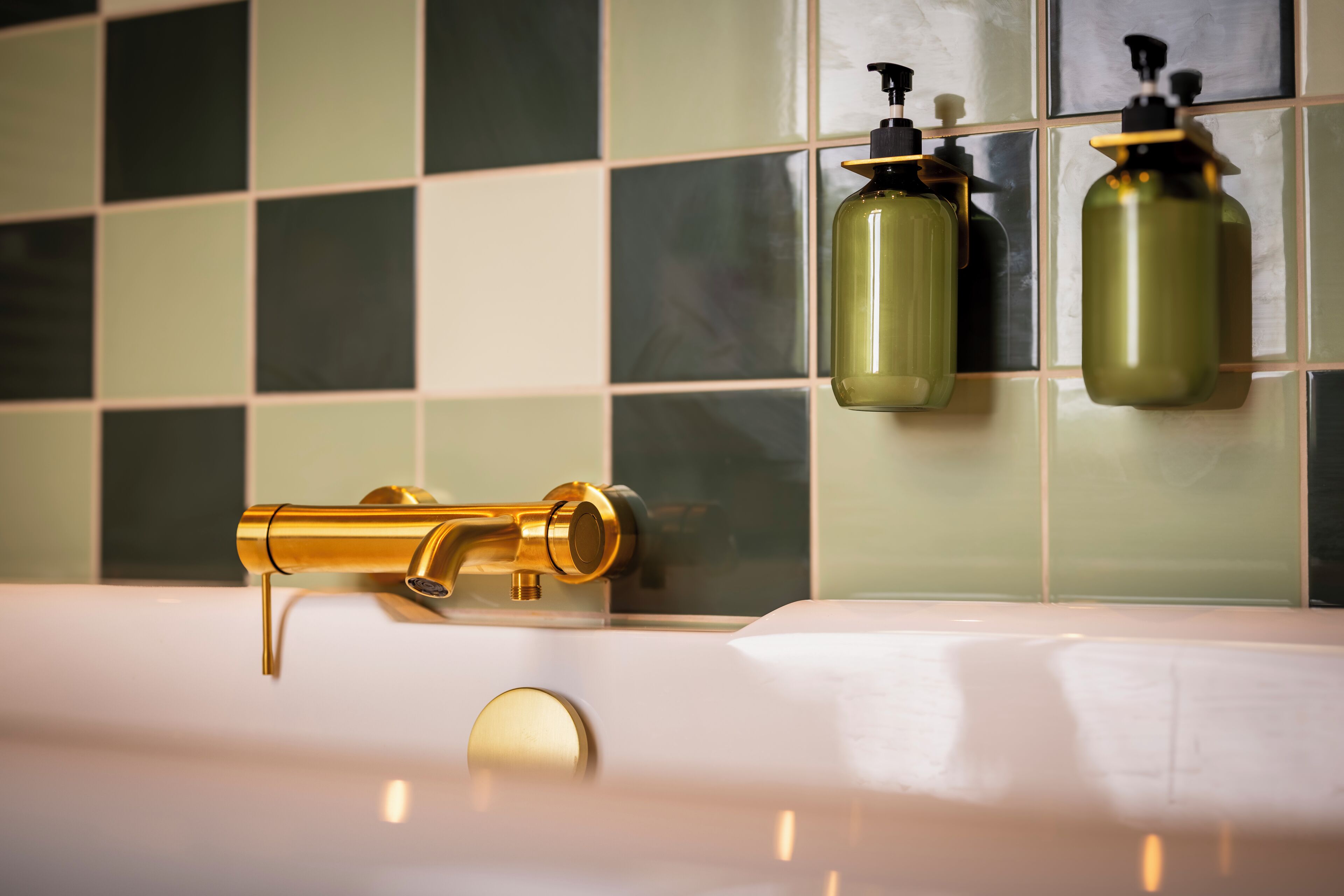 Une salle de bain avec un robinet doré, deux distributeurs de savon et un mur recouvert de carreaux.