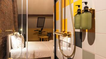 Quarto de hotel com uma cama, um separador de vidro, banheiro com azulejos amarelos e brancos, e duas garrafas de shampoo.