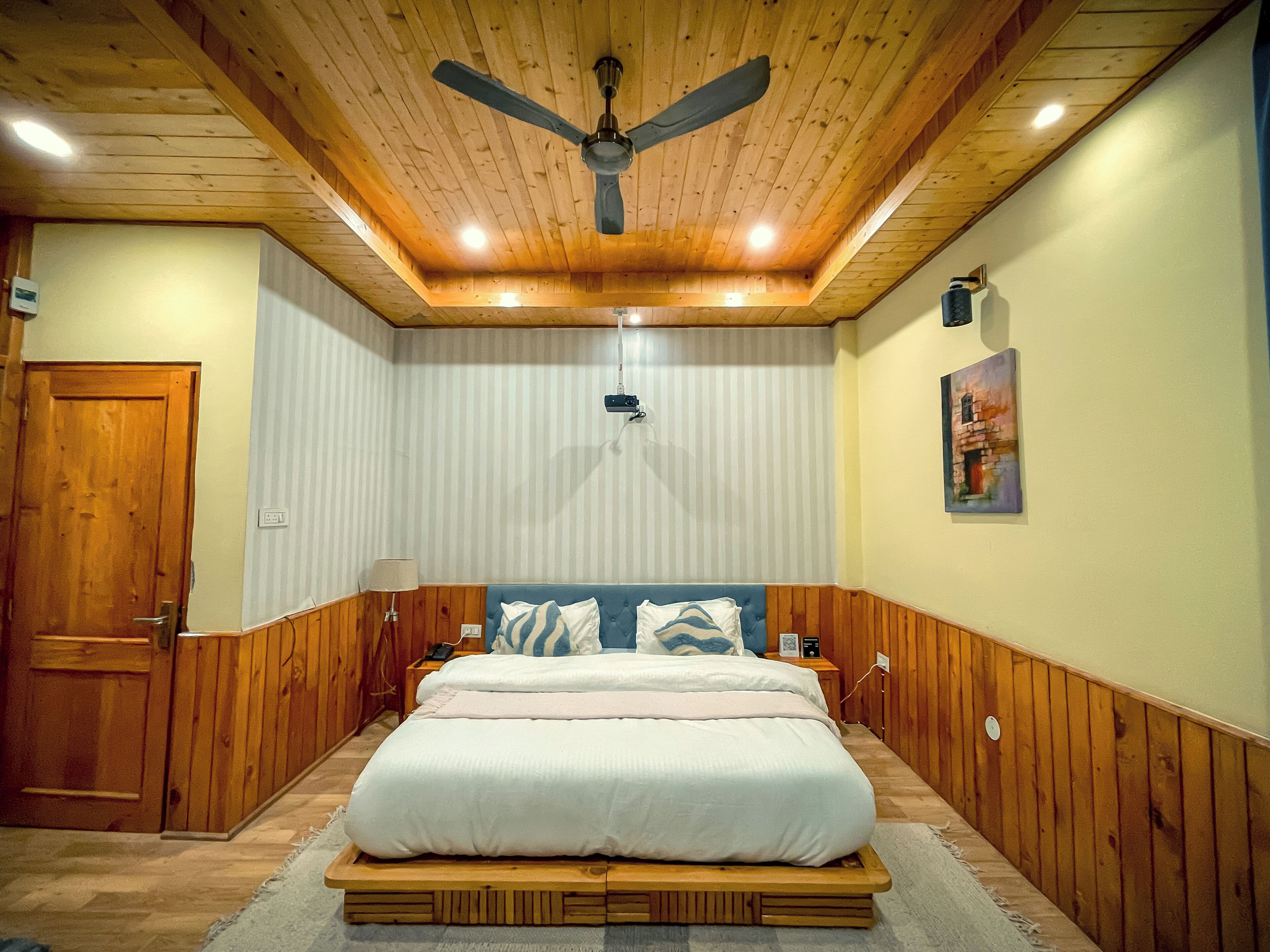 Una habitación con techo de madera, ventilador de techo, cama con ropa de cama blanca, mesita de noche y un proyector montado en la pared.