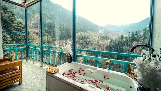 A bathroom with a bathtub, a wooden bench, and a view of a mountainous landscape through large windows. - The Elevia Hotels (Banjar)