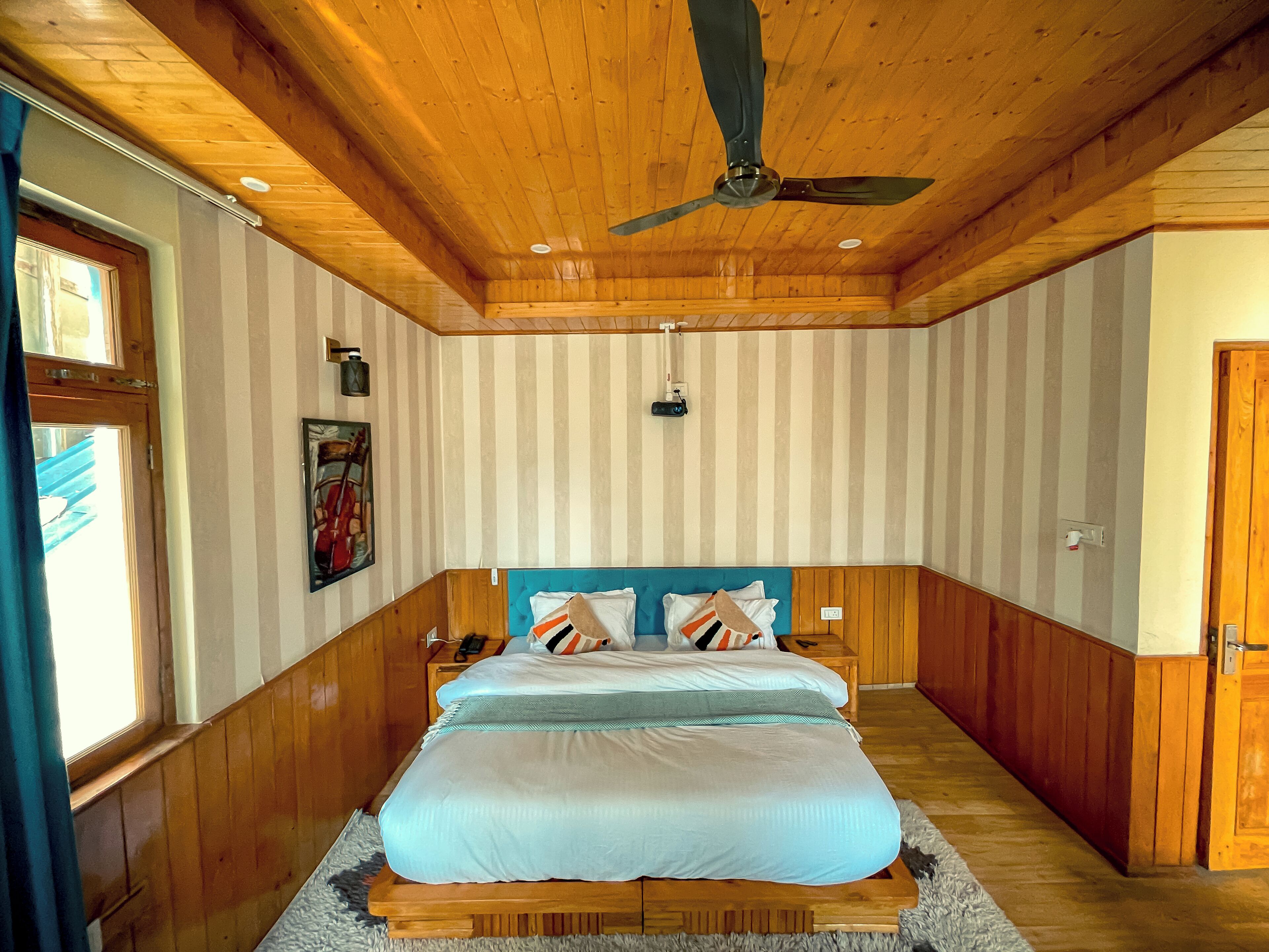 A bedroom with a wooden ceiling, a bed with a blue headboard, striped wallpaper, and a ceiling fan.