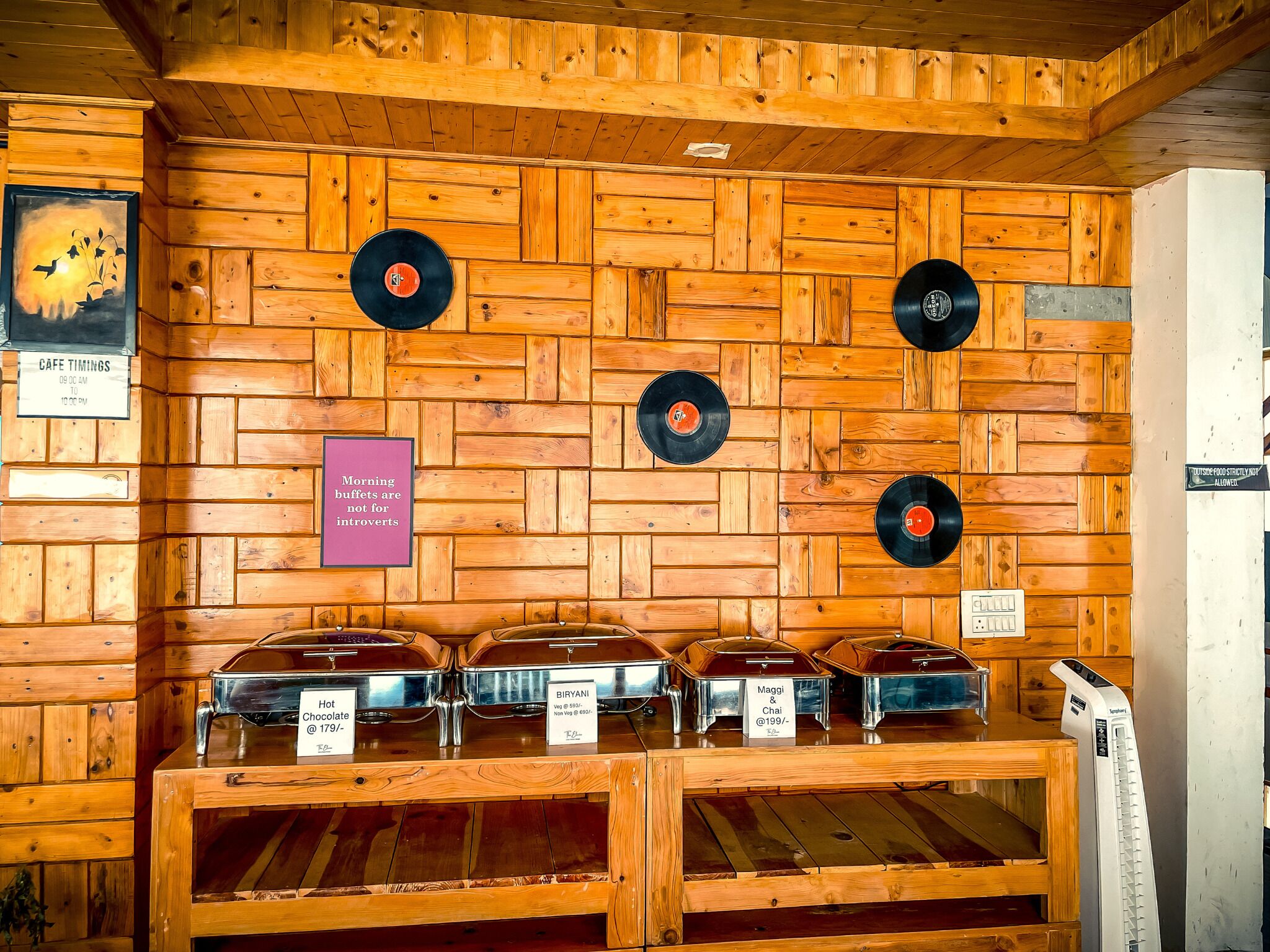 A wooden wall with a record player, a sign for cafe timings, and buffet containers labeled with food items.