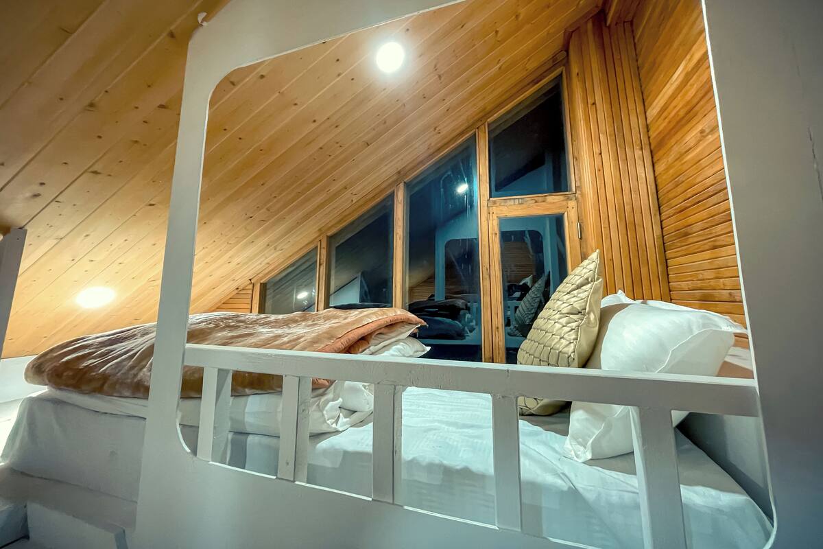 Quarto com teto inclinado, paredes de madeira, cama com roupa de cama branca e janela com persianas de madeira.