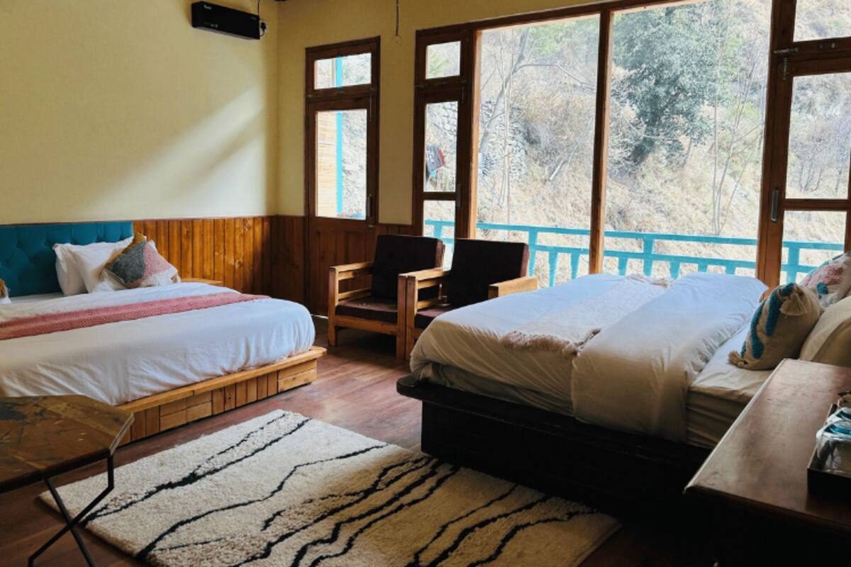 Quarto com duas camas, uma escrivaninha de madeira e vista para árvores através de grandes janelas.