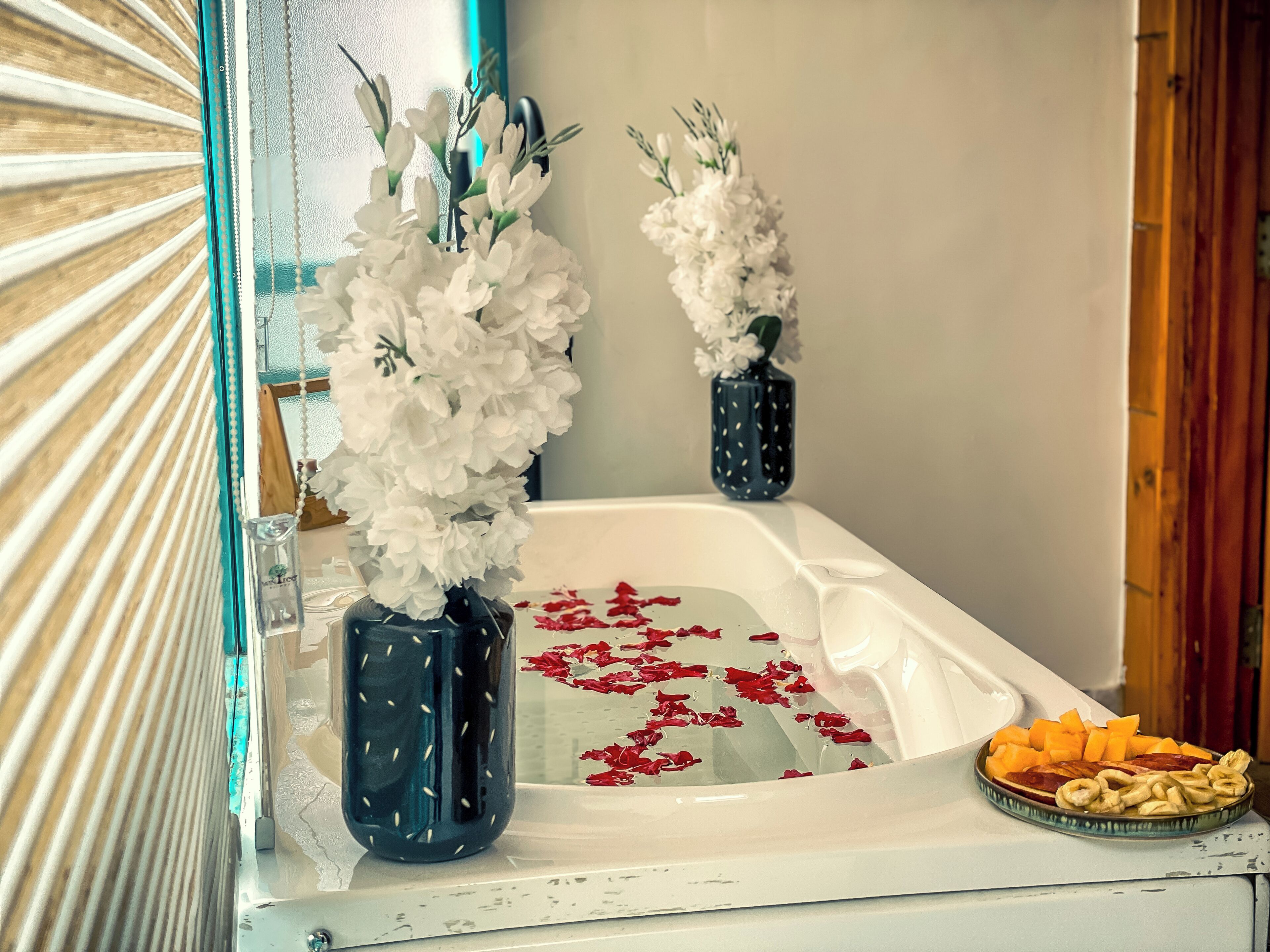 A bathtub with white flowers in vases, red petals, and a plate of snacks.