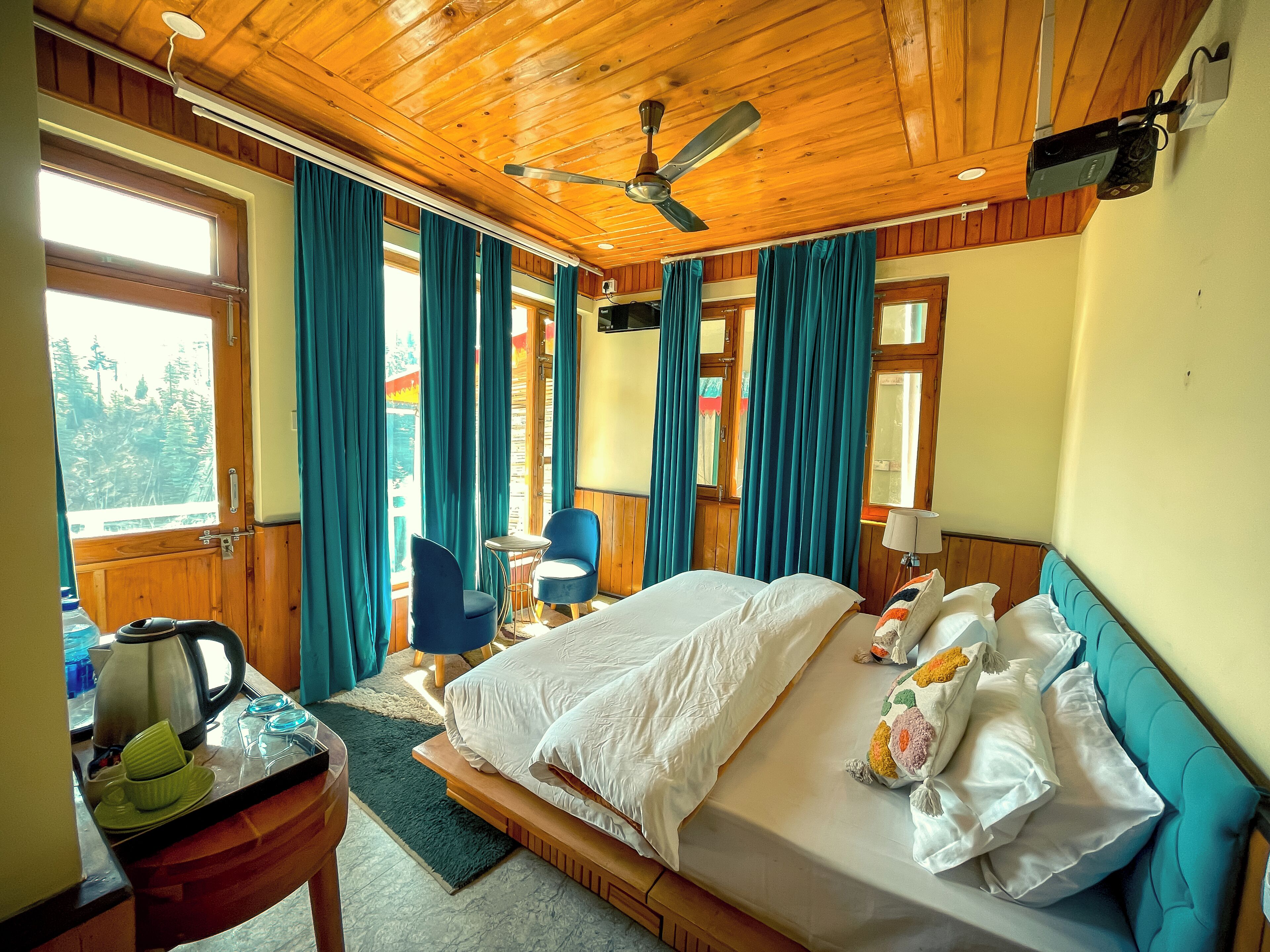 A hotel room with a wooden ceiling, a bed with white linens, a small table with a kettle, and blue chairs.