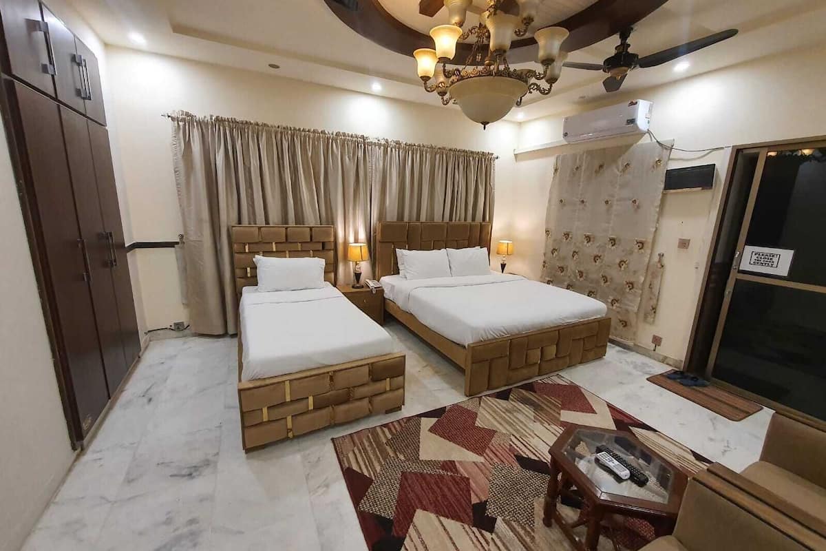 A hotel room with two beds, a ceiling fan, a chandelier, a patterned rug, and a small table with a remote control.