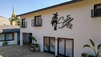 Welcome to Siesta, a contemporary building with a flat roof and decorative railings, surrounded by lush greenery.