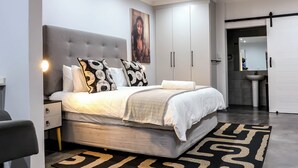 Een moderne slaapkamer met een grijs gewatteerd hoofdeinde, een groot bed met gekleurde kussens, een nachtkastje en een inloopkast met schuifdeuren.